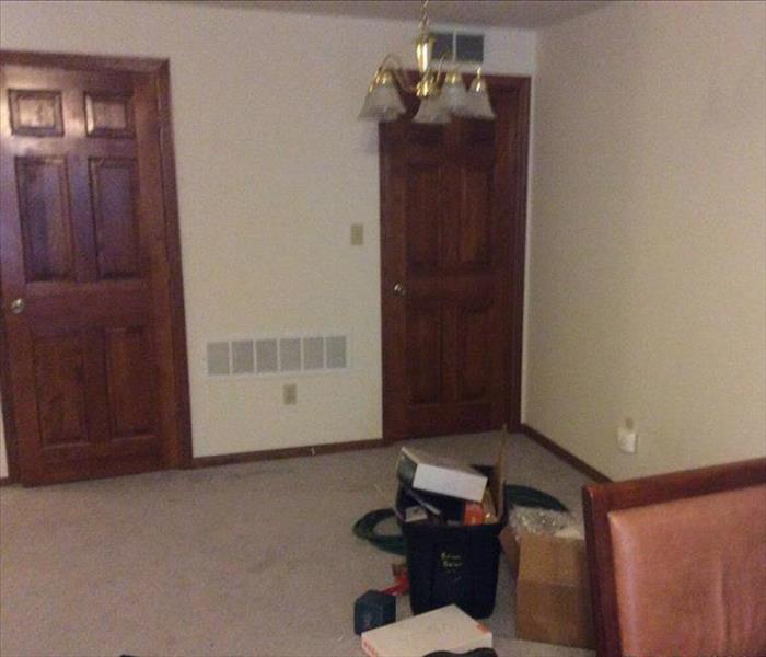 A small carpeted room with two exterior doors has a couple boxes and chairs and water damage to the flooring